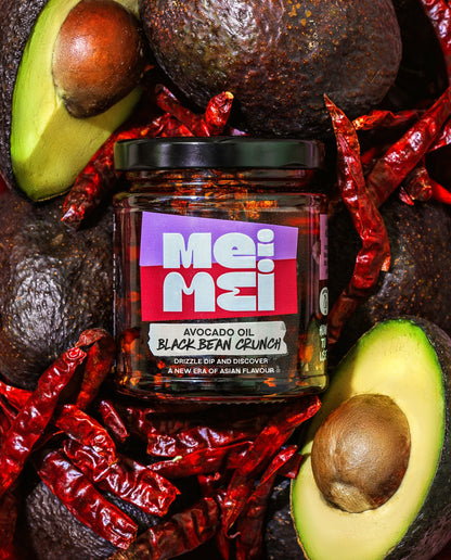 Jar of Mei Mei Avocado Oil Black Bean Crunch surrounded by avocados and red chili peppers.