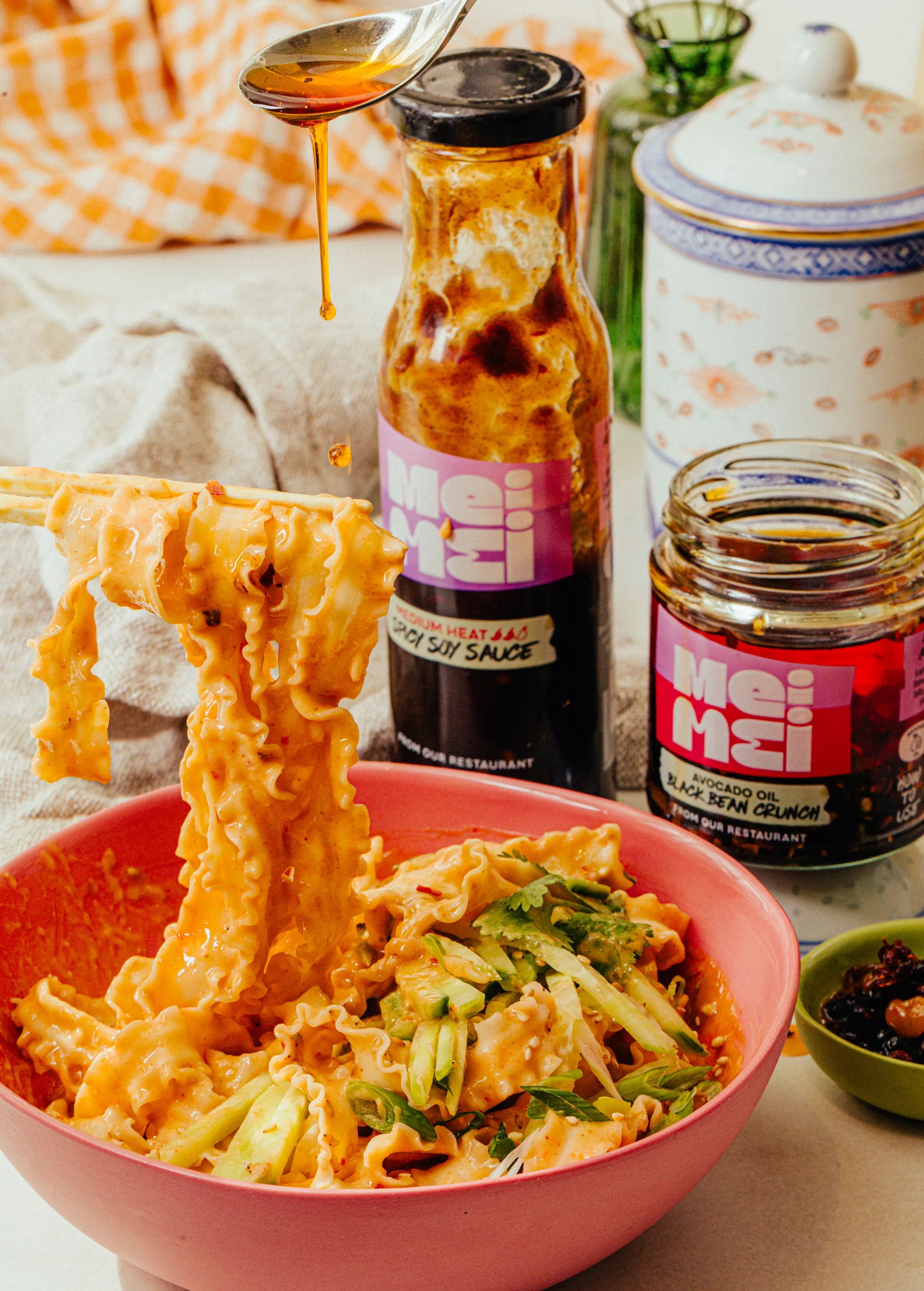 Noodles with vegetables in a pink bowl, Mei Mei chili oil sauce bottle in the background