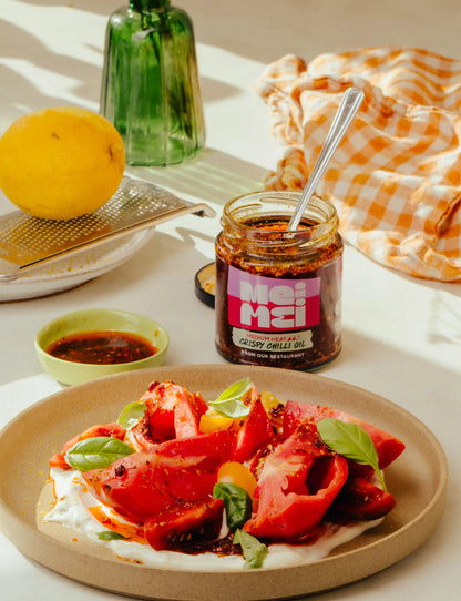 Mei Mei crispy chilli oil jar on a table with a dish of tomatoes and basil.