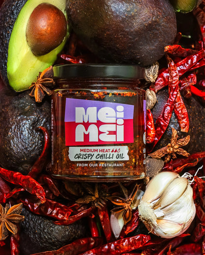 Jar of Mei Mei Crispy Chilli Oil surrounded by avocados, red chilies, and garlic