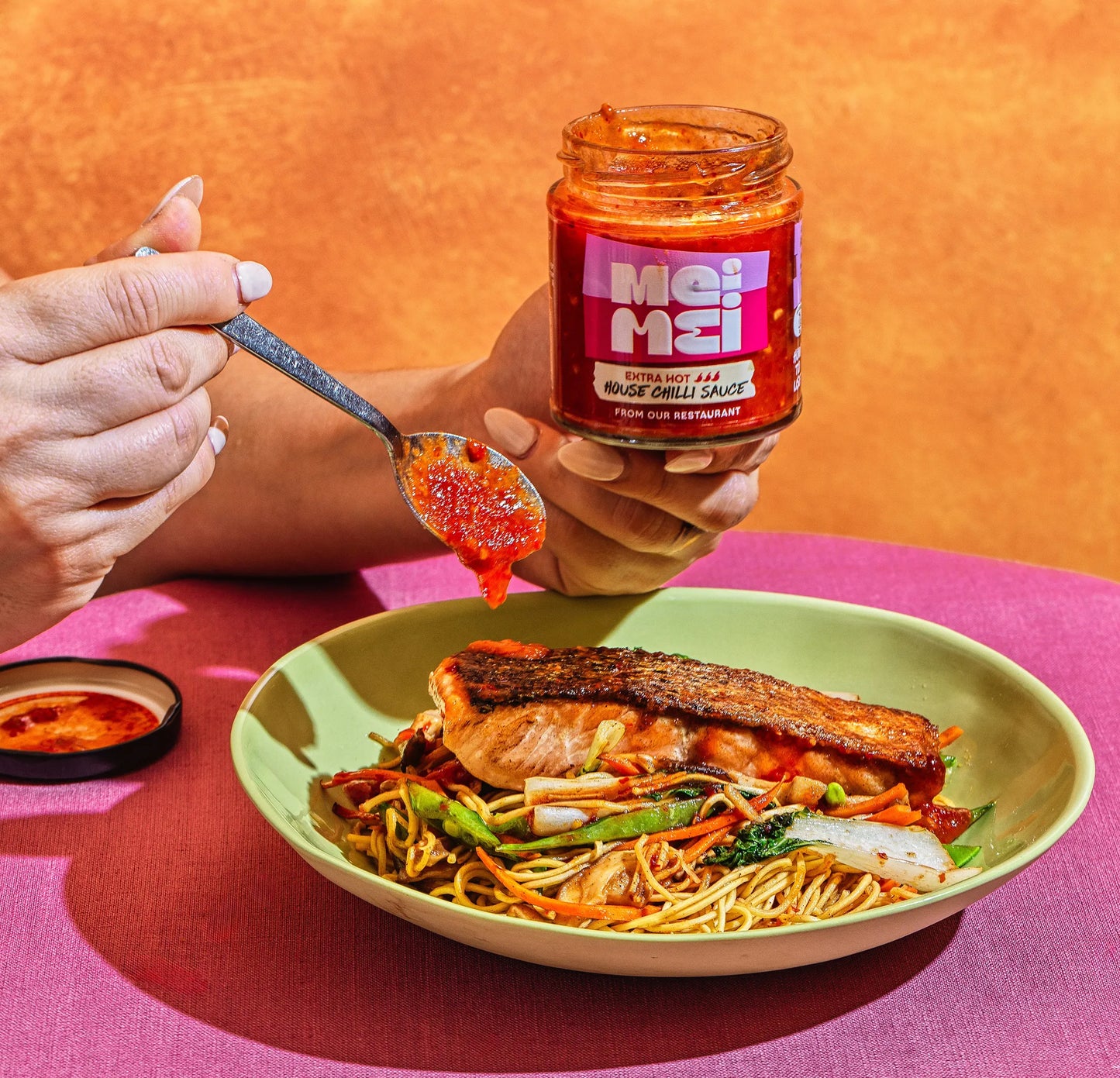 Person holding a jar of Mei Mei chilli sauce over a bowl of noodles with a spoonful of sauce.
