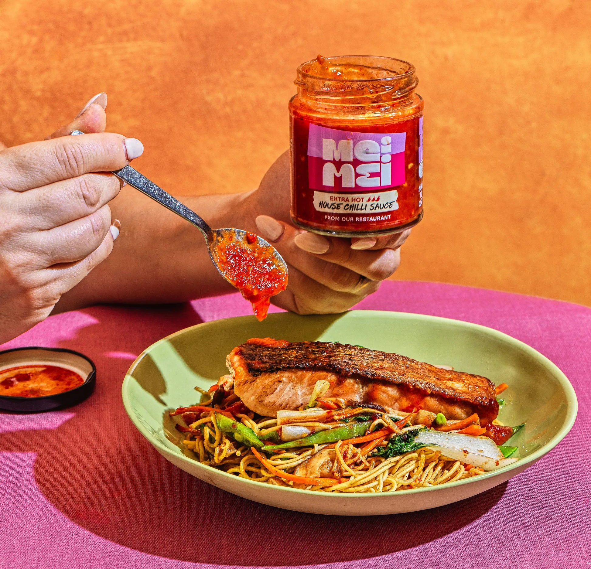 Person holding a jar of Mei Mei chilli sauce over a bowl of noodles with a spoonful of sauce.